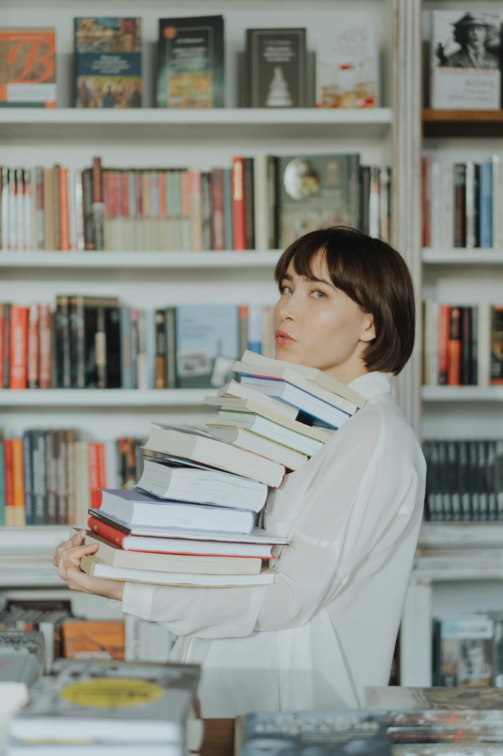A woman in a bookstore holding a large stack of books, wearing a white shirt.