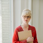 Blonde woman in red shawl holding clipboard by window, creating a serene academic vibe.